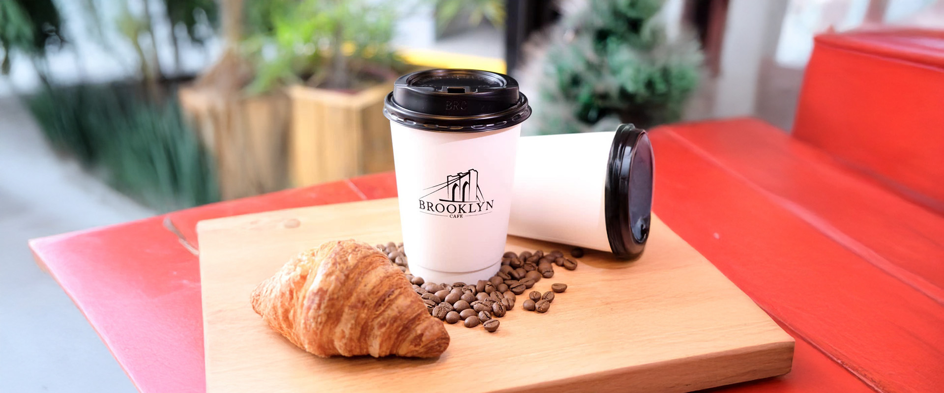 A Brooklyn Cafe takeaway cup and a croissant on a wooden board with coffee beans