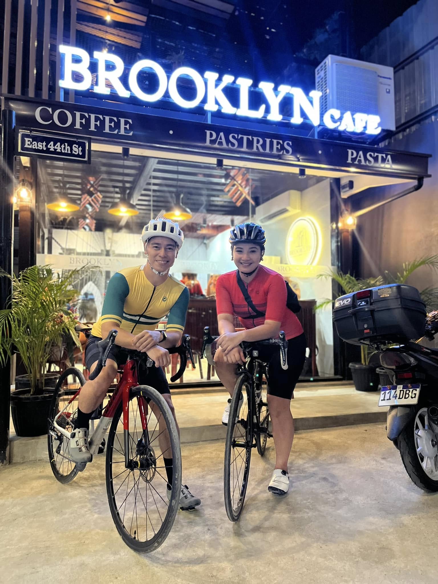 Two cyclists with their road bikes in front of the lit Brooklyn Cafe sign